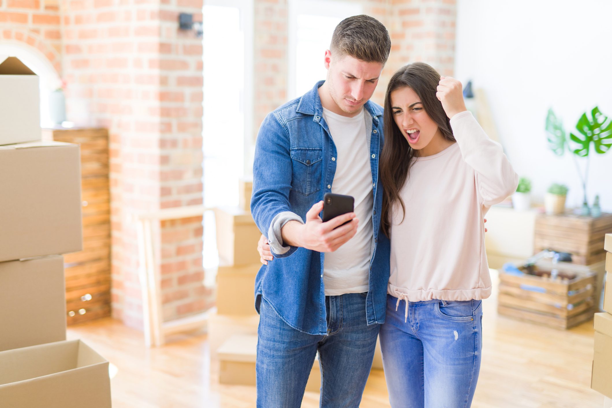 Beautiful young couple moving to a new house using smartphone annoyed and frustrated shouting with anger, crazy and yelling with raised hand, anger concept