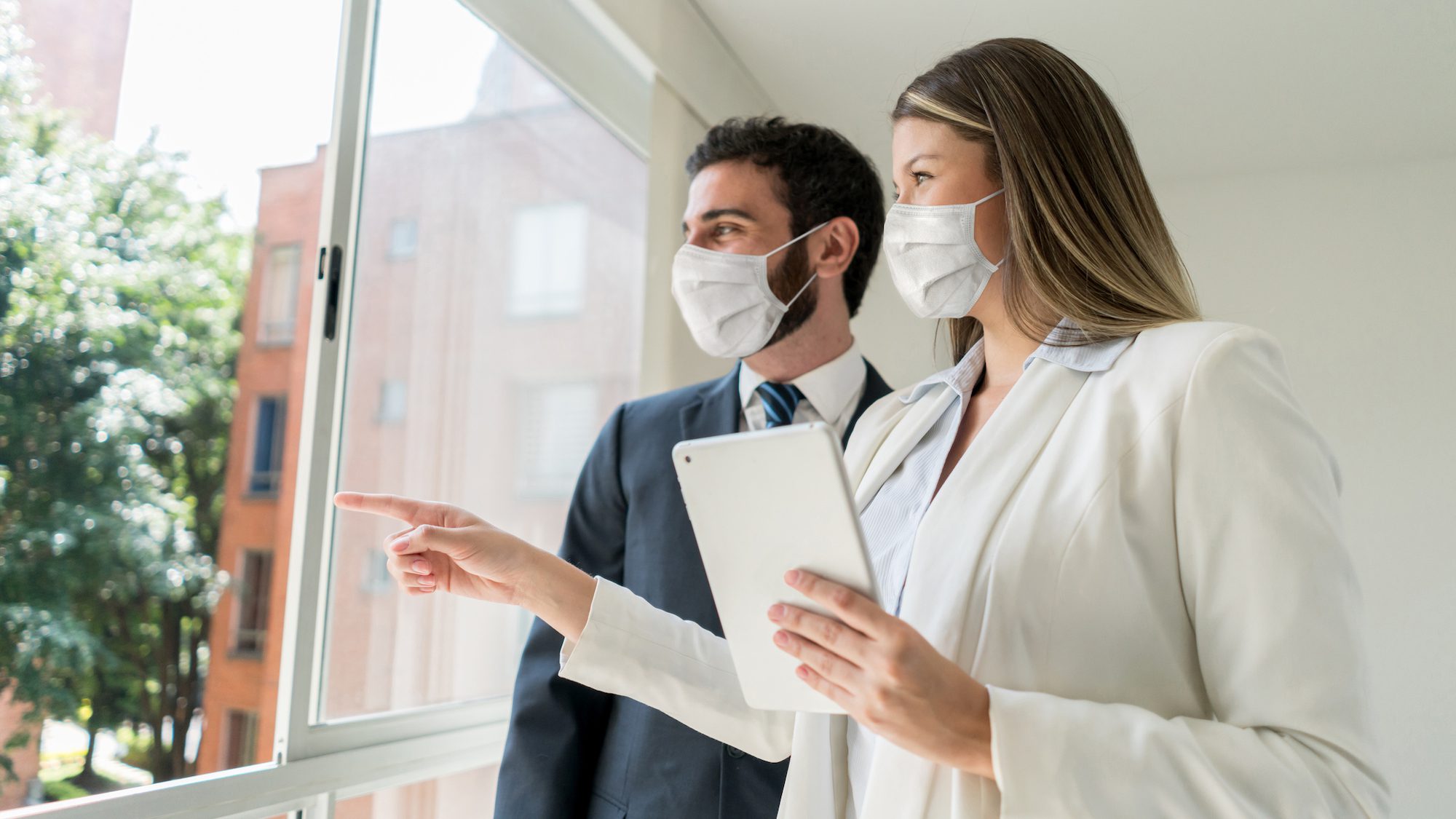 Beautiful real estate agent pointing at the view to customer both looking very cheerful but wearing protective face masks